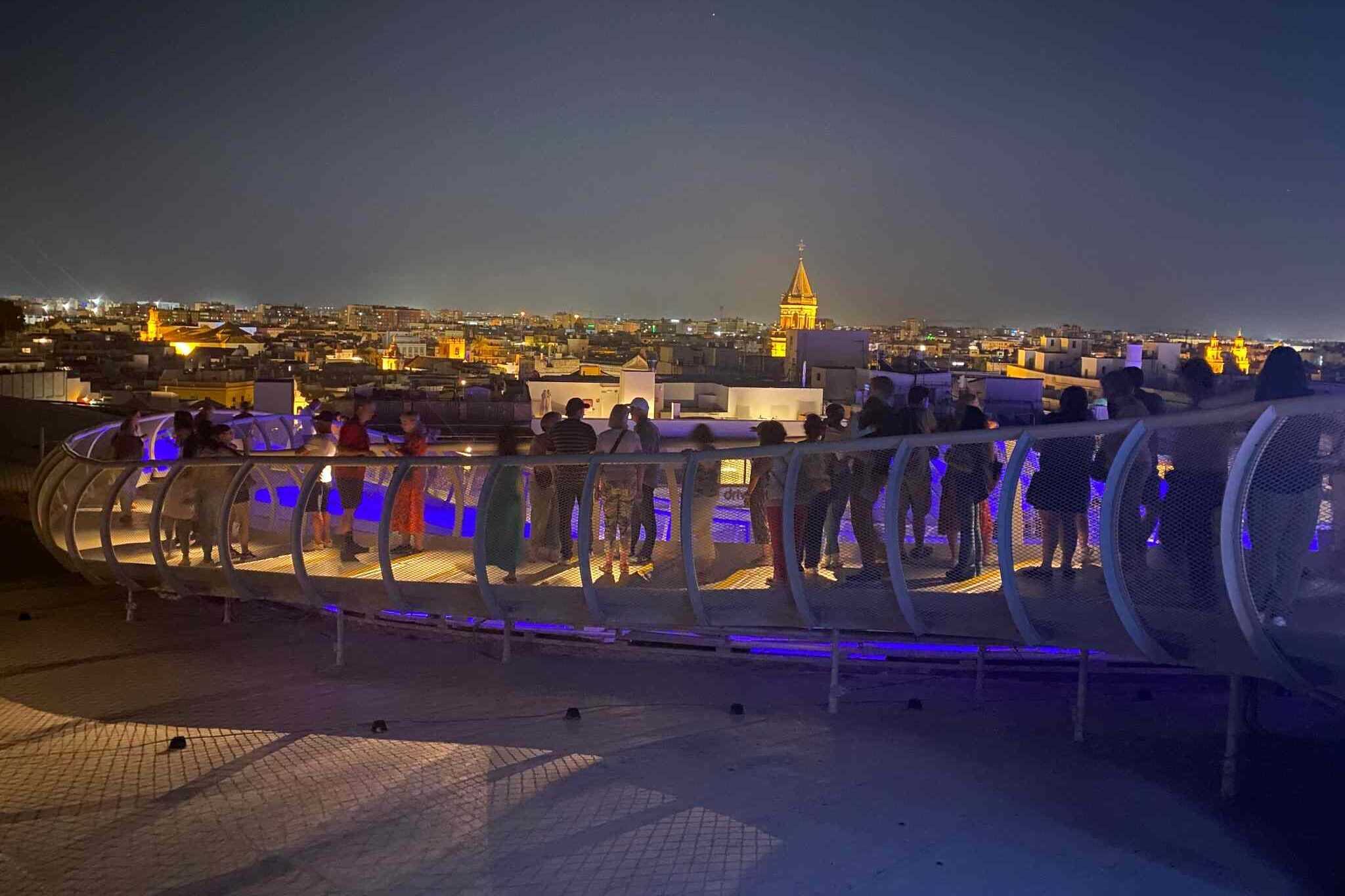 People walking along the illuminated walkway of the Metropol Parasol in Seville with city rooftops in the background